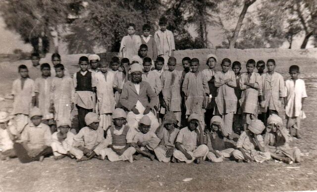 Schoolchildren seated and standing in rows outdoors with their teachers.