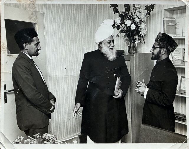 Hazrat Khalifatul Masih III standing between two men, smiling at the man on the right.