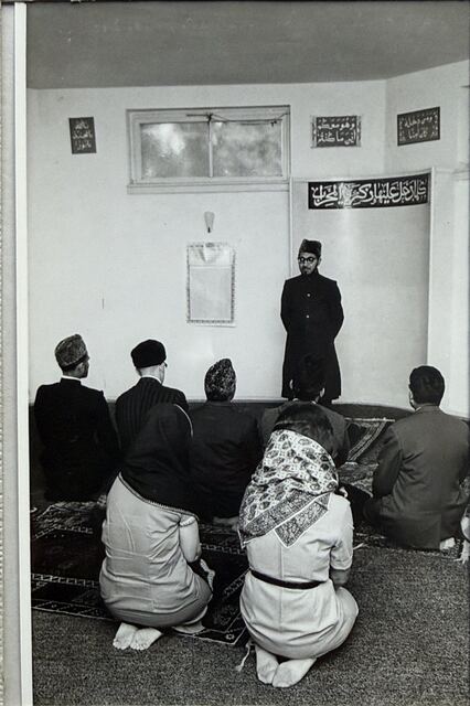 A man standing at the front of the prayer hall, people seated before him.